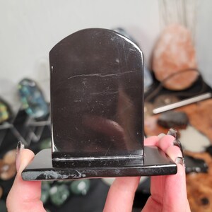 May include: A black, rectangular bookend with a rounded top. The bookend is made of a glossy, dark material and has a smooth surface. The base is also rectangular and the same dark color. The bookend is held by a person.