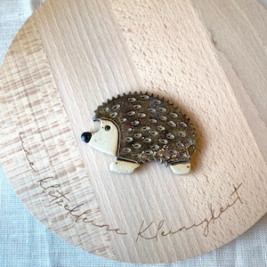 Könnte beinhalten: Eine Keramik-Igel-Figur mit strukturiertem, stacheligem Rücken, die auf einem hellen Holzschneidebrett liegt. Das Brett hat eine runde Form und ein natürliches Holzmaserungsmuster. Der Text "eine klitzekleine Kleinigkeit" ist auf dem Brett geschrieben.