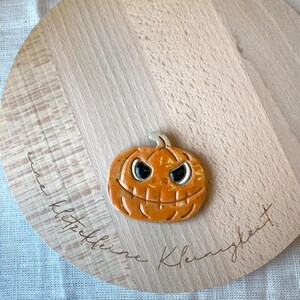 Könnte beinhalten: Ein Keramik-Kürbis mit einem geschnitzten Gesicht und einem zahnigen Grinsen. Der Kürbis ist orange mit schwarzen Augen und einem weißen Mund. Der Kürbis liegt auf einem Holzbrett mit dem eingravierten Text "eine kleine Kleinigkeit".