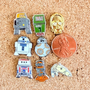 May include: A collection of Star Wars character enamel pins, including R2-D2, C-3PO, and BB-8, in various colors. A penny is included for size reference. The pins are displayed on a corkboard surface.