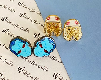 May include: Two pairs of enamel pin earrings. One pair features a blue and red character with a white outline. The other pair features a gold character with a white helmet and red accents.