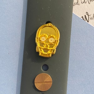 May include: A gold-colored watch band with a small, gold-colored C-3PO character charm. The band has a black background and a small, round, gold-colored screw.