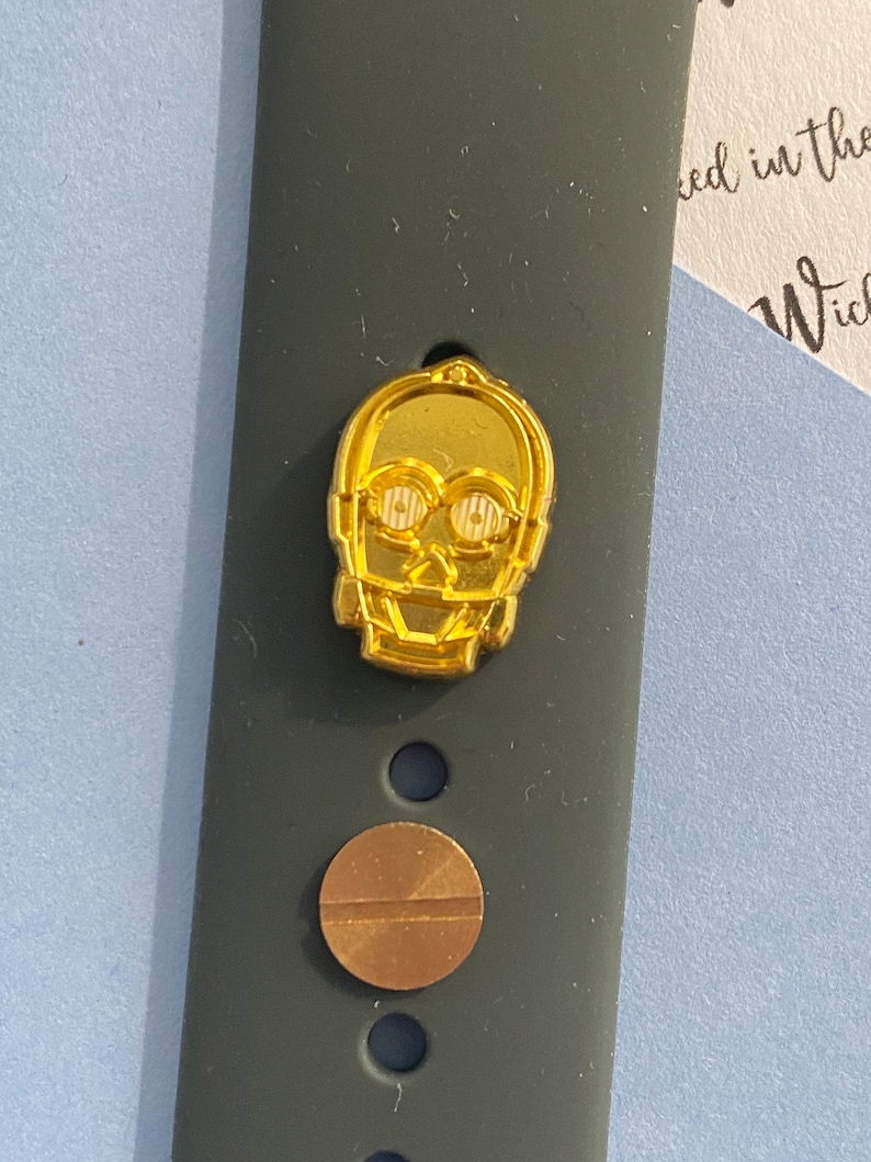 May include: A gold-colored watch band with a small, gold-colored C-3PO character charm. The band has a black background and a small, round, gold-colored screw.