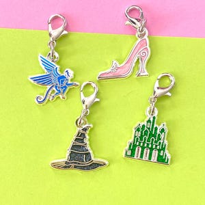 May include: Four silver charm pendants with different designs. The charms are a blue and silver winged monkey, a pink glass slipper, a green castle, and a black and silver witch's hat.