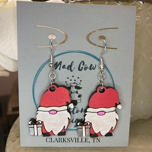 May include: A pair of wooden gnome earrings with red hats and white beards. Each gnome holds a silver gift box. The earrings hang from silver hooks. The background includes the text "Mad Cow" and "Clarksville, TN".