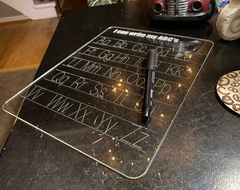 May include: A clear plastic dry-erase board with the alphabet printed on it. The board is on a black surface with a black marker next to it. The text on the board reads "I can write my ABC's".