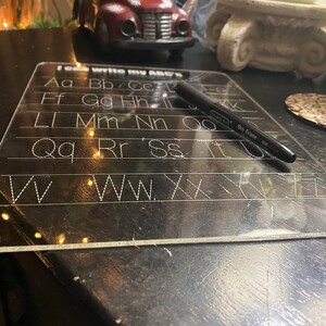 Puede incluir: Una pizarra transparente de borrado en seco de plástico con líneas punteadas para trazar el alfabeto. La pizarra dice "I can write my ABC's" e incluye un marcador de borrado en seco negro.