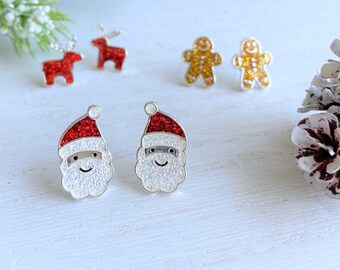 Puede incluir: Un par de pendientes de Navidad plateados con caras de Papá Noel con gorros rojos y barbas blancas. Los pendientes están decorados con purpurina.