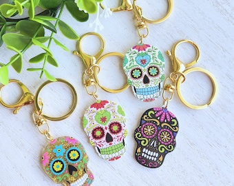 May include: Four gold keychains with colorful sugar skull charms. Each skull has a different design with flowers and swirls. The keychains are on a white wooden surface.