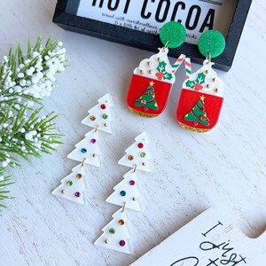 Puede incluir: Pendientes navideños festivos. Un par presenta tazas de cacao rojas con crema batida y un árbol de Navidad. El otro par tiene formas de árbol blancas con gemas de colores. Un letrero dice "Hot Cocoa".