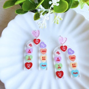 May include: A pair of rainbow candy heart earrings. Each earring is shaped like a candy heart and features a different Valentine's Day message on each heart. The messages include "Hug Me", "Love You", "Cutie", "Be Mine", "XOXO", "Best Buds", "Te Amo", "Kiss Me", and "Mwah".