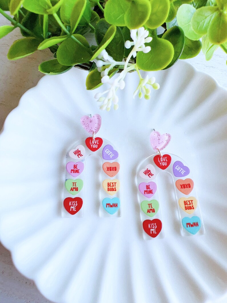 May include: A pair of rainbow candy heart earrings. Each earring is shaped like a candy heart and features a different Valentine's Day message on each heart. The messages include "Hug Me", "Love You", "Cutie", "Be Mine", "XOXO", "Best Buds", "Te Amo", "Kiss Me", and "Mwah".