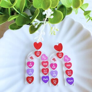 May include: A pair of Valentine's Day earrings with a candy heart design. Each earring features a clear acrylic base with a series of colorful heart charms. The charms have various messages, including "Hug Me", "Love You", "Cutie", "Be Mine", "XOXO", "Best Buds", "Te Amo", "Kiss Me", and "Mwah".
