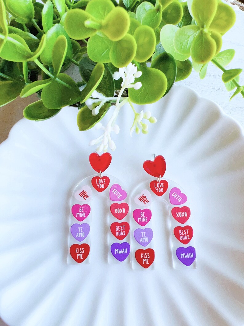 May include: A pair of Valentine's Day earrings with a candy heart design. Each earring features a clear acrylic base with a series of colorful heart charms. The charms have various messages, including "Hug Me", "Love You", "Cutie", "Be Mine", "XOXO", "Best Buds", "Te Amo", "Kiss Me", and "Mwah".