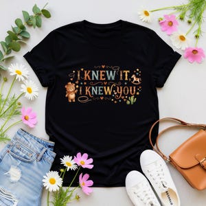 Puede incluir: Una camiseta negra de manga corta con un estampado gr&aacute;fico que incluye el texto "I KNEW IT, I KNEW YOU". El dise&ntilde;o muestra un oso de peluche marr&oacute;n con sombrero de vaquero, un caballito balanc&iacute;n, estrellas, corazones y un cactus.