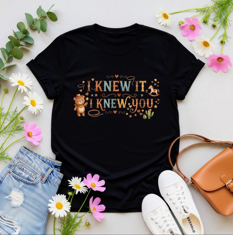 Puede incluir: Una camiseta negra de manga corta con un estampado gr&aacute;fico que incluye el texto "I KNEW IT, I KNEW YOU". El dise&ntilde;o muestra un oso de peluche marr&oacute;n con sombrero de vaquero, un caballito balanc&iacute;n, estrellas, corazones y un cactus.