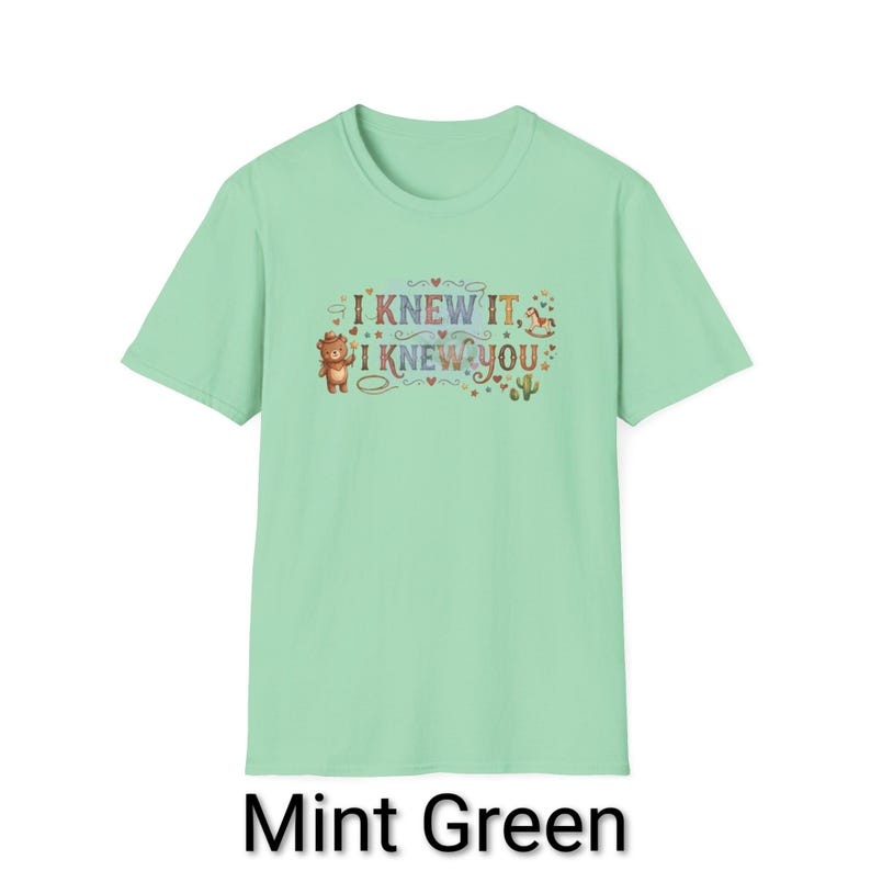 Puede incluir: Una camiseta de manga corta color verde menta con un estampado gr&aacute;fico que incluye el texto I KNEW IT, I KNEW YOU. El dise&ntilde;o presenta un oso marr&oacute;n, un peque&ntilde;o caballito de madera, un cactus y varios corazones y estrellas decorativas.