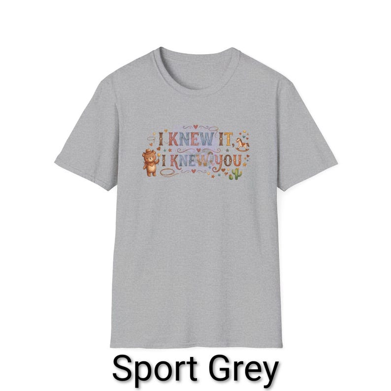 Puede incluir: Una camiseta de manga corta color gris deportivo con un estampado gr&aacute;fico que incluye el texto I KNEW IT, I KNEW YOU. El dise&ntilde;o presenta un peque&ntilde;o oso marr&oacute;n, un caballito de madera, un cactus y varios corazones y estrellas de colores.