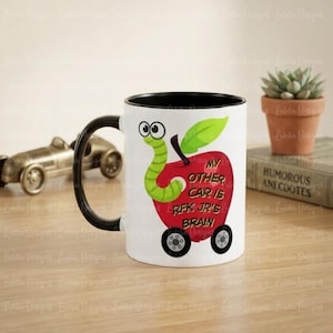 May include: A white ceramic mug with a black handle and rim, featuring a graphic of a red apple with wheels and a green worm. The apple has text that reads: MY OTHER CAR IS RFK JR'S BRAIN. The design includes a green leaf on top of the apple.