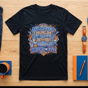 Op de afbeelding: Een zwart T-shirt met korte mouwen en een grafische print met de tekst MY MAYOR MUSLIM MY BAGELS JEWISH MY SATURDAY NIGHT LIVE KNICKS IN FIVE, omringd door basketbal-illustraties, samen met een blauwe spijkerbroek, een riem, een pet en een horloge.