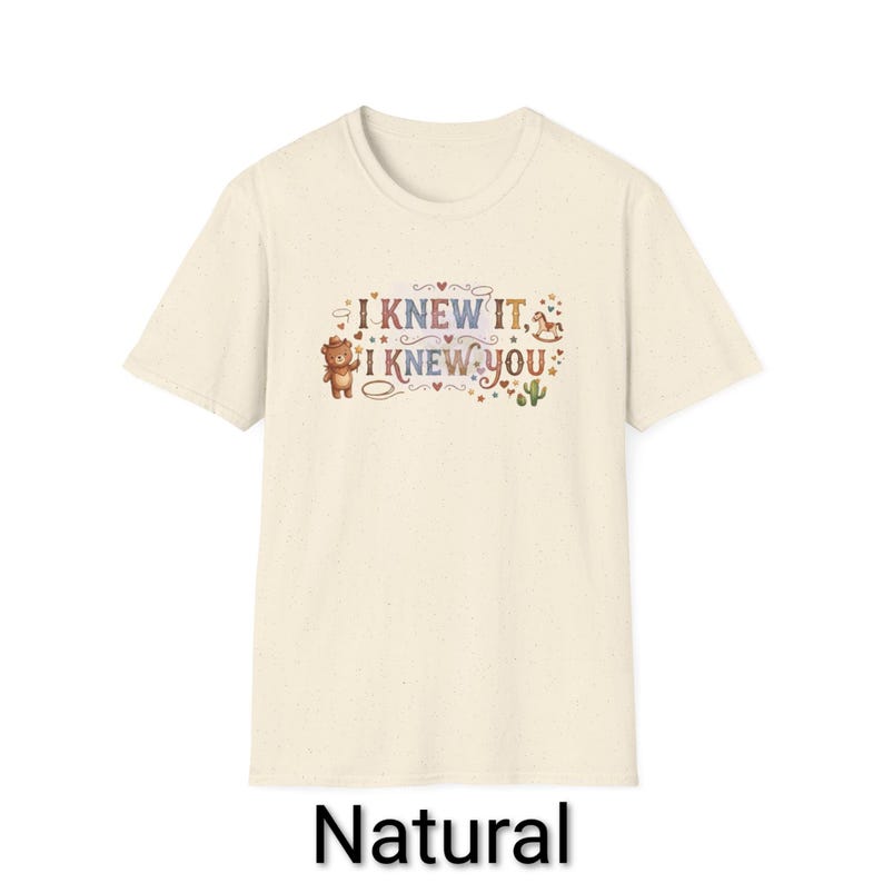 Puede incluir: Una camiseta de manga corta de color natural con un estampado gr&aacute;fico que incluye el texto "I KNEW IT, I KNEW YOU". El dise&ntilde;o presenta un peque&ntilde;o oso marr&oacute;n, un caballito de madera, un cactus y varias estrellas y corazones en tonos tierra.