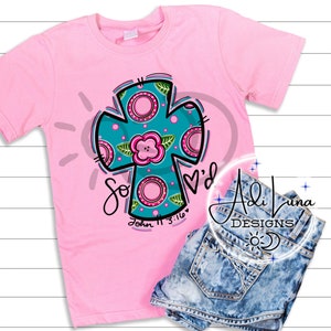May include: Pink t-shirt with a teal cross design featuring pink flowers and white polka dots. The cross has the words "So Loved" and "John 11:3:16" written below it.