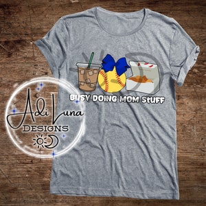 May include: Gray t-shirt with a graphic design of a coffee cup, a softball with a blue bow, and a box of chicken nuggets. The text "Busy Doing Mom Stuff" is printed below the graphic.