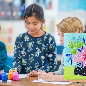 May include: Three children are painting at a table. One child is painting a canvas with a floral design. The canvas is on an easel. The other two children are painting on paper. There are paint bottles on the table.