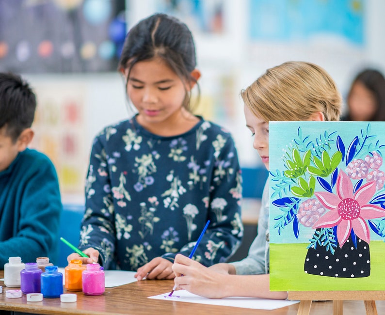 May include: Three children are painting at a table. One child is painting a canvas with a floral design. The canvas is on an easel. The other two children are painting on paper. There are paint bottles on the table.