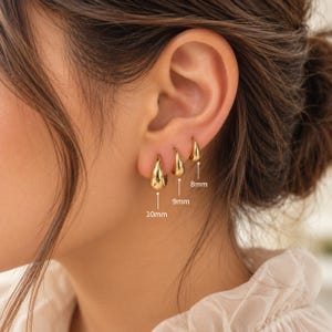Può includere: Tre orecchini a cerchio dorati a forma di goccia, disposti in fila sul lobo dell'orecchio. Gli orecchini sono etichettati con le rispettive dimensioni: 10mm, 9mm e 8mm, indicando il diametro di ogni singolo orecchino a cerchio del set.