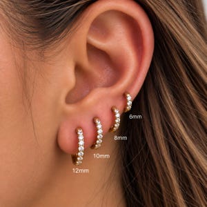 May include: Four gold hoop earrings with clear stones arranged in a row on an earlobe. The hoops are labelled with sizes 6mm, 8mm, 10mm, and 12mm. Each earring features a circular band set with small, sparkling stones, showcasing a graduated size progression.