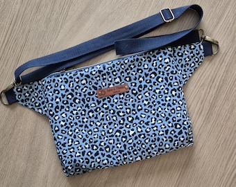 May include: A blue fanny pack with a leopard print pattern in black and white. The bag has a navy blue adjustable strap and a zipper closure. A small brown leather tag is attached to the front.