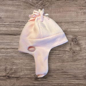 May include: A pink fleece hat with a pom-pom on top and a face opening. The hat has a long flap that can be pulled down to cover the neck.