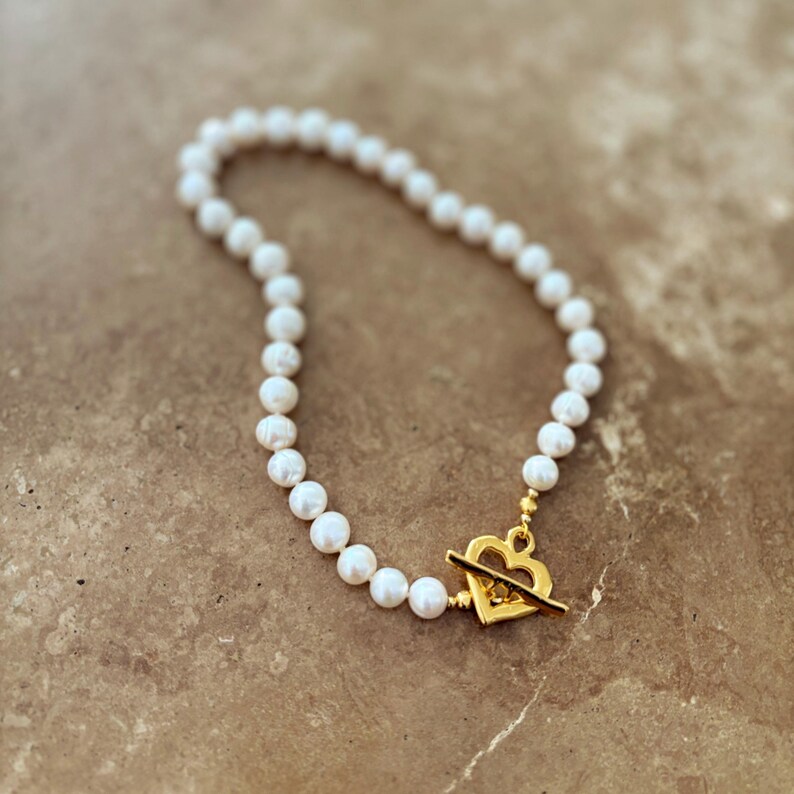 May include: A necklace featuring a strand of white pearls arranged in a circular shape. The necklace is secured with a gold-toned metal toggle clasp that is shaped like an open heart, with a horizontal bar passing through the center of the heart.