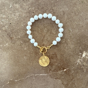 May include: A bracelet featuring a strand of white, round, lustrous pearls connected to a gold-toned toggle clasp. A circular gold-toned coin charm with embossed detailing hangs from the centre of the toggle mechanism, completing the design.