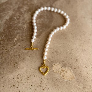 May include: A necklace featuring a strand of white pearls with a gold-toned toggle clasp and a gold-toned open heart-shaped pendant. The pearls are arranged in a circular shape, and the metal components have a textured, slightly irregular finish.