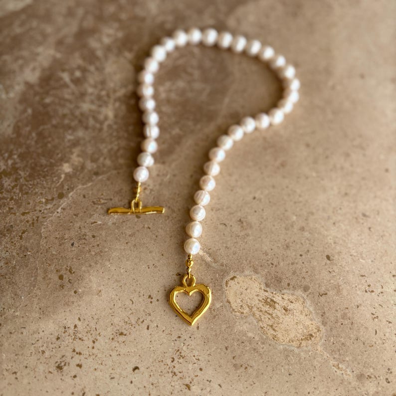May include: A necklace featuring a strand of white pearls with a gold-toned toggle clasp and a gold-toned open heart-shaped pendant. The pearls are arranged in a circular shape, and the metal components have a textured, slightly irregular finish.