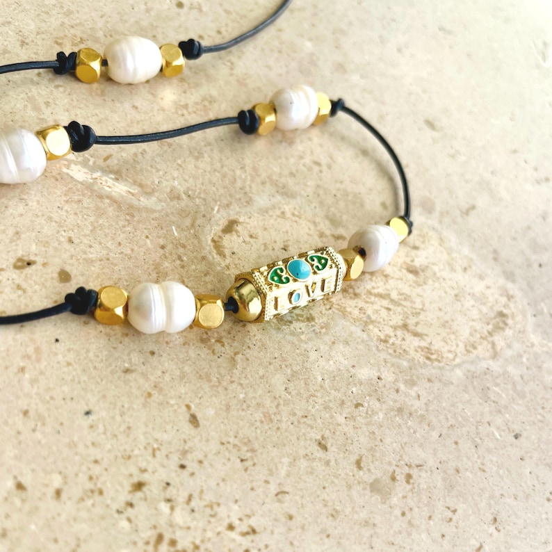 May include: Close-up of a necklace featuring white pearls, gold-colored beads, and a gold-colored rectangular charm with the word "LOVE" and turquoise accents. The necklace is strung on a black cord.