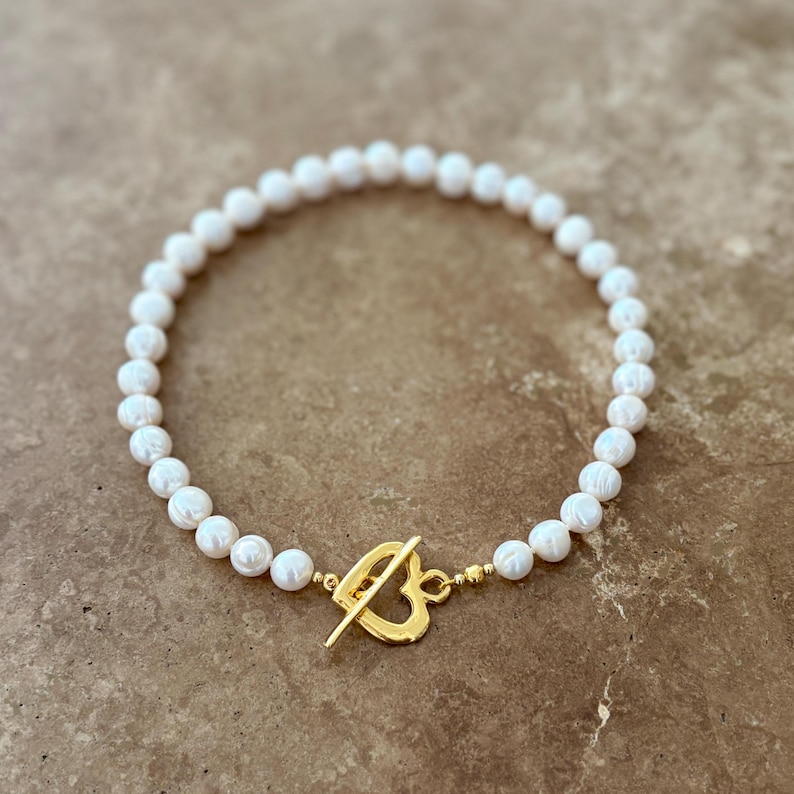 May include: A necklace composed of a single strand of white, round pearls. The necklace features a gold-toned metal toggle clasp shaped like an open heart, positioned at the front, connecting the two ends of the pearl strand with small gold beads.