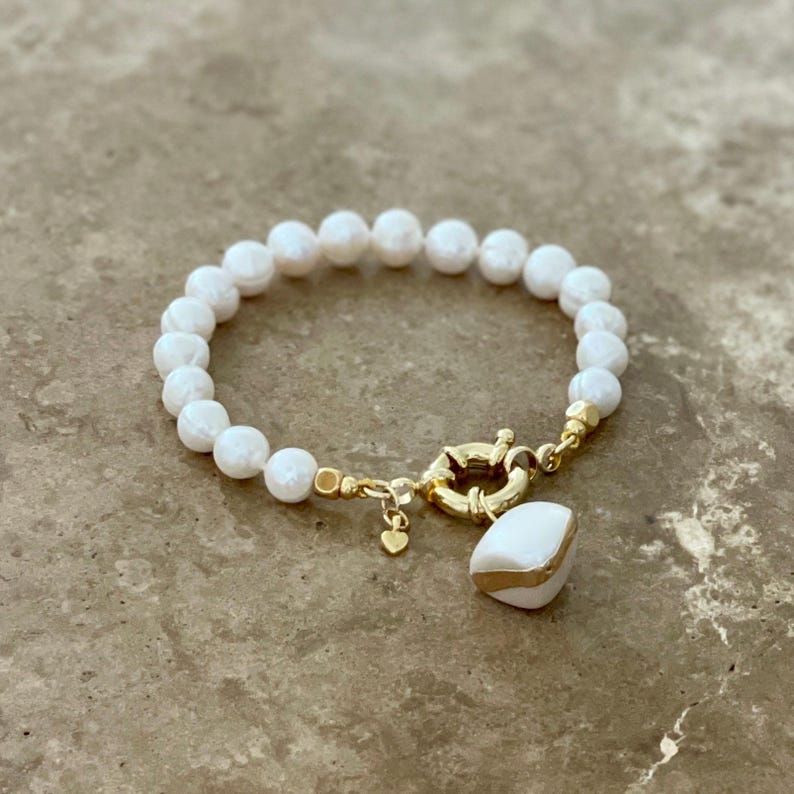 K&ouml;nnte beinhalten: Ein wei&szlig;es Perlenarmband mit einem goldenen Verschluss und einem wei&szlig;en und goldenen Anh&auml;nger. Das Armband liegt auf einer hellbraunen Oberfl&auml;che.