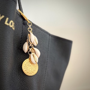 Op de afbeelding: Een goudkleurige bedel met een munt- en schelpmotief hangt aan een zwarte draagtas. De bedel heeft een ronde, reliëf munt en verschillende kleine, crèmekleurige schelpen met gouden accenten. De tas heeft gouden letters.