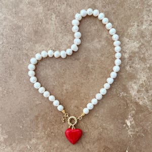 Peut inclure: Un collier compos&eacute; de perles d'eau douce blanches de forme irr&eacute;guli&egrave;re dispos&eacute;es en cercle. Le collier pr&eacute;sente un fermoir circulaire dor&eacute; &agrave; la base, auquel est suspendu un pendentif lisse en forme de c&oelig;ur rouge vif.