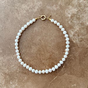 Peut inclure: Un collier compos&eacute; de nombreuses perles blanches de forme organique et arrondie, enfil&eacute;es en un motif circulaire. Le collier pr&eacute;sente un fermoir &agrave; ressort circulaire en m&eacute;tal dor&eacute; sur le dessus, reliant les deux extr&eacute;mit&eacute;s du rang de perles.
