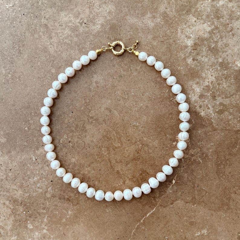 Peut inclure: Un collier compos&eacute; de nombreuses perles blanches de forme organique et arrondie, enfil&eacute;es en un motif circulaire. Le collier pr&eacute;sente un fermoir &agrave; ressort circulaire en m&eacute;tal dor&eacute; sur le dessus, reliant les deux extr&eacute;mit&eacute;s du rang de perles.