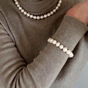May include: A beige turtleneck sweater paired with a matching set of jewelry consisting of a single-strand pearl necklace and a coordinating pearl bracelet. The pearls are round, white, and uniform in size, creating a classic and cohesive look.