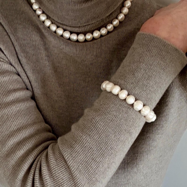 May include: A beige turtleneck sweater paired with a matching set of jewelry consisting of a single-strand pearl necklace and a coordinating pearl bracelet. The pearls are round, white, and uniform in size, creating a classic and cohesive look.