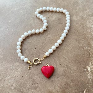 Peut inclure: Un collier compos&eacute; d'un rang de perles d'eau douce blanches reli&eacute;es par un fermoir circulaire dor&eacute;. Un pendentif en forme de c&oelig;ur rouge lisse et brillant est suspendu au fermoir, qui comporte &eacute;galement une petite breloque &eacute;toile.