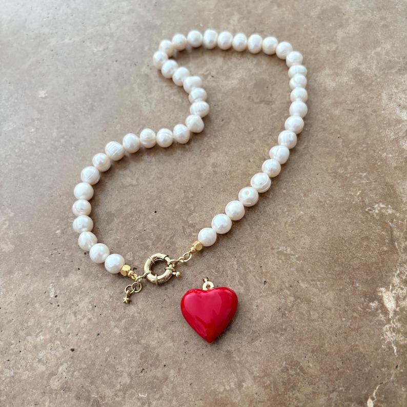 Peut inclure: Un collier compos&eacute; d'un rang de perles d'eau douce blanches reli&eacute;es par un fermoir circulaire dor&eacute;. Un pendentif en forme de c&oelig;ur rouge lisse et brillant est suspendu au fermoir, qui comporte &eacute;galement une petite breloque &eacute;toile.