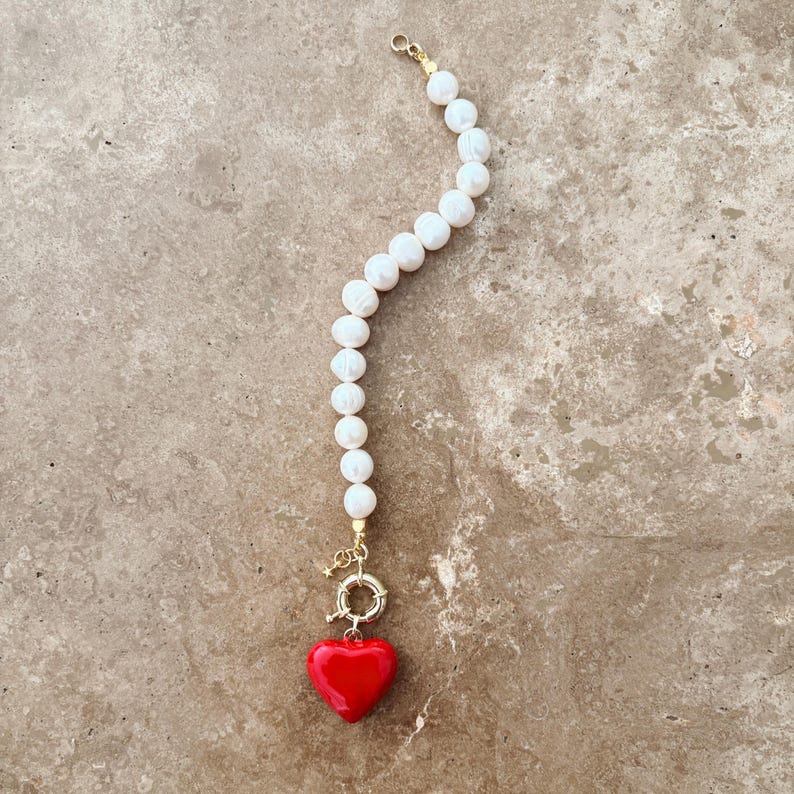 May include: A bracelet featuring a strand of white, irregularly shaped pearls arranged in a curved line. The bracelet includes a gold-toned metal toggle clasp and a single, smooth, bright red heart-shaped charm suspended from the clasp.