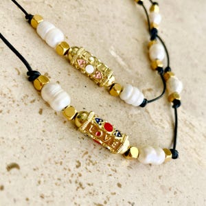 May include: A close-up of a necklace featuring white pearl beads, gold-colored accents, and black cord. The necklace has gold-colored rectangular beads with the word "LOVE" and colorful accents. The necklace is on a beige surface.